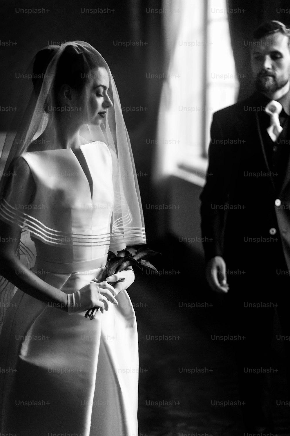 elegant gorgeous bride gently looking under veil at stylish groom standing at window light, holding calla bouquet. wedding couple in retro style. black white photo