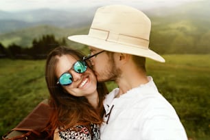 stylish hipster man kissing his woman on top of mountains and smiling in summer, travel together concept, space for text