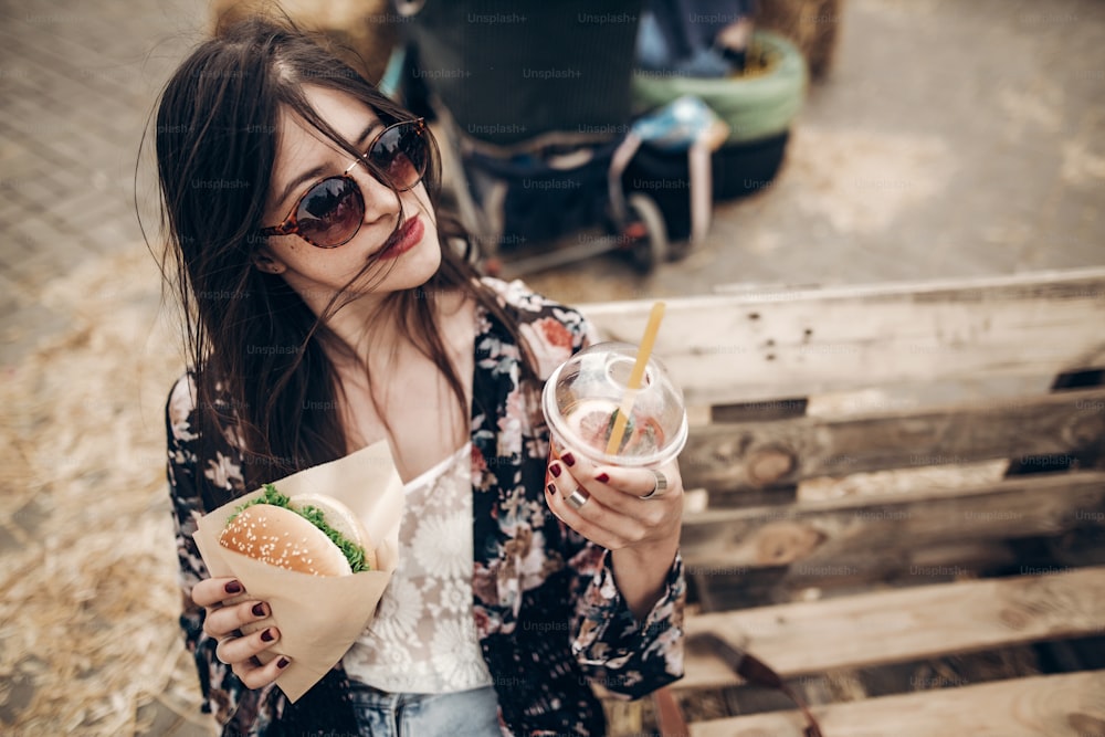 Elegante mujer hipster sosteniendo jugosa hamburguesa y limonada. Chica boho con hamburguesa y bebida sonriendo en el festival de comida callejera. verano. Viajes de vacaciones de verano. Espacio para el texto