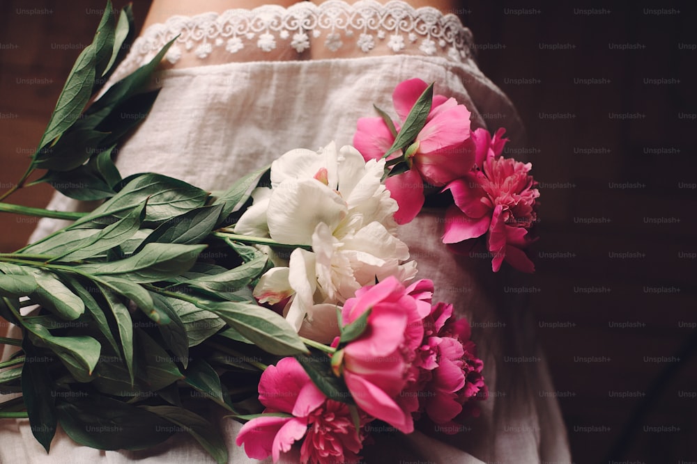 흰색 보헤미안 드레스를 입은 보헤미안 소녀의 다리에 사랑스러운 분홍색 모란, 상위 뷰. 텍스트 공간. 아침 방에 아름 다운 꽃과 함께 앉아 세련 된 힙 스터 여자입니다. 분위기 있는 관능적인 순간
