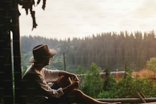 Hipster-Mann sitzt auf der Veranda des Holzhauses und schaut abends auf den Wald, ruhiger Moment, Sommerurlaubskonzept, Platz für Text