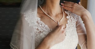 Hermosa novia sosteniendo un costoso collar de plata con perlas en el cuello. Mujer en vestido blanco con adornos florales de encaje, preparativos de la mañana nupcial. joyería con estilo. Foto de tocador