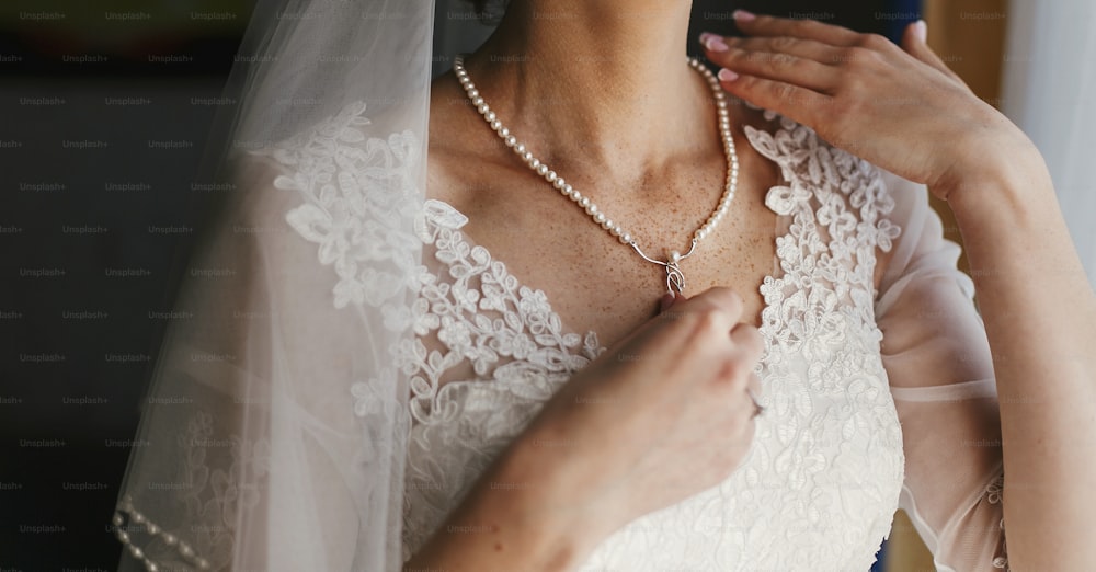 beautiful bride holding expensive silver necklace with pearls on neck. woman in white gown with lace floral ornaments, bridal morning preparations. stylish jewelry. boudoir photo