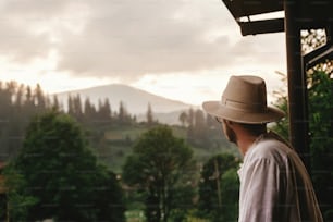 Hipster-Mann auf der Veranda des Holzhauses mit Blick auf die Berge im abendlichen Sonnenuntergang, ruhiger entspannender Moment, Sommerurlaubskonzept, Platz für Text