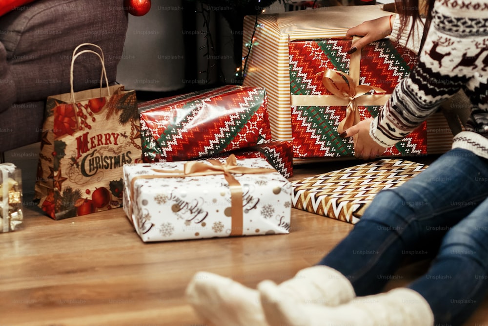 woman stacking christmas presents under tree. gift ideas. girl in sweater with deers and wearing santa hat in festive room with lights. seasonal greetings concept. joyful moment