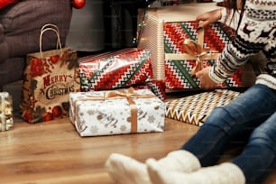 donna che impila i regali di natale sotto l'albero. Idee regalo. ragazza in maglione con cervi e che indossa cappello da babbo natale nella stanza festiva con le luci. concetto di saluti stagionali. momento gioioso