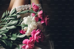 beautiful pink peonies on legs of boho girl in white bohemian dress, top view. space for text. stylish hipster woman sitting with flowers in moody morning. atmospheric sensual moment