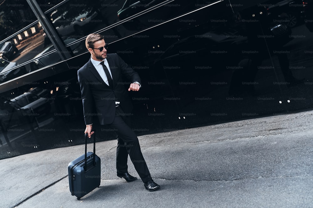 Full length of young man in full suit pulling luggage while walking outdoors