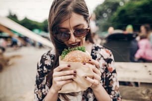 mulher hipster elegante segurando hambúrguer suculento e comendo. menina boho com hambúrguer sorrindo no festival de comida de rua. Verão. viagens de férias de verão. espaço para texto