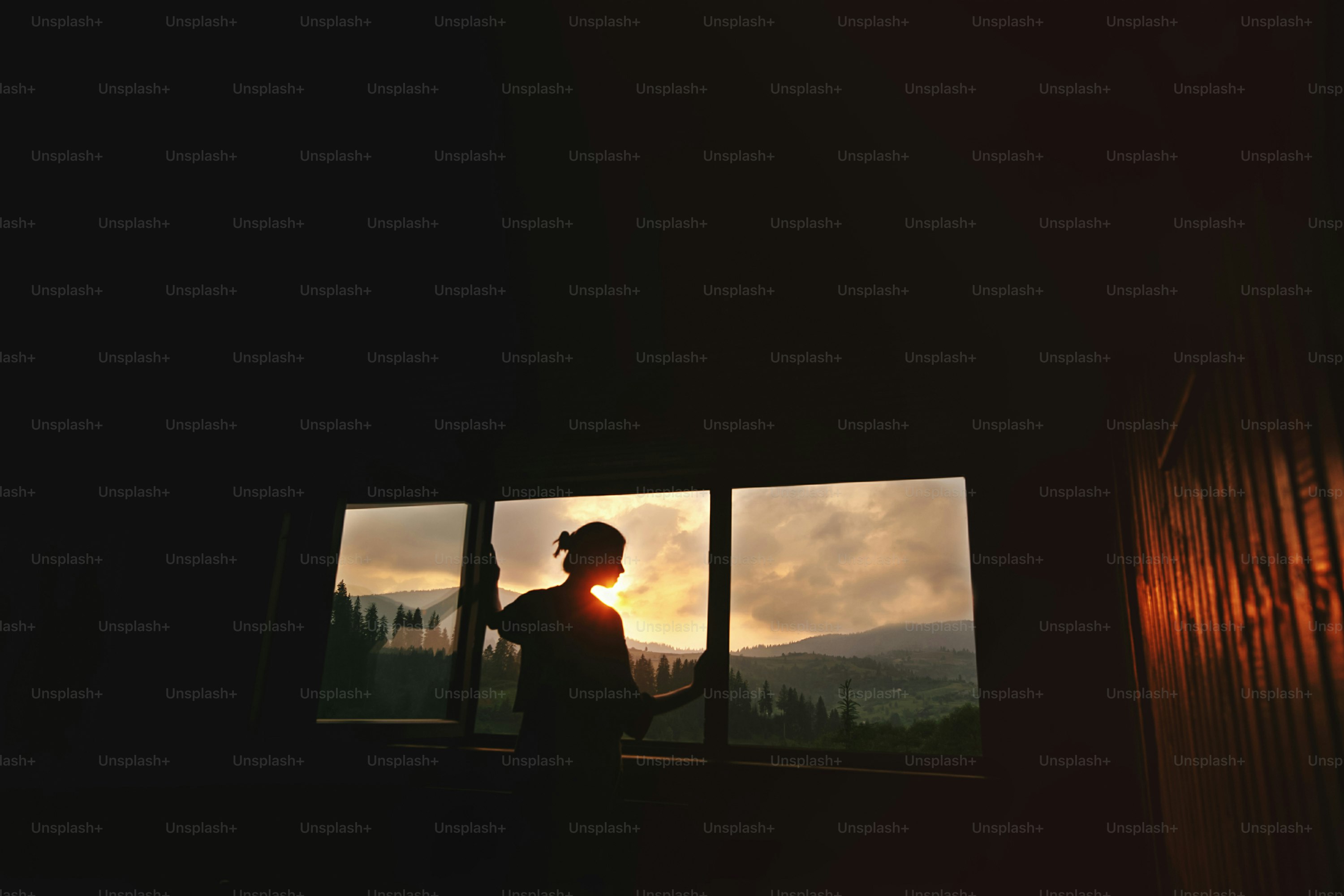 Silueta de mujer en la luz del sol en la ventana de madera de la cabaña con vista a la puesta del sol en las montañas, momento tranquilo del concepto de relajación, espacio para el texto