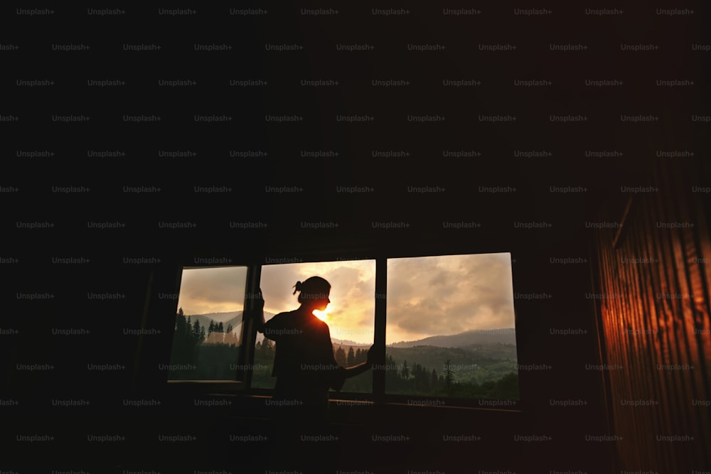 silhouette di donna al sole in finestra di legno cottage con vista sul tramonto in montagna, momento tranquillo di concetto di relax, spazio per il testo