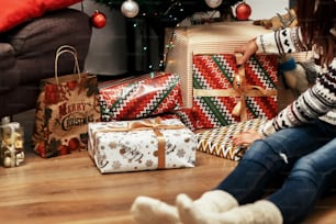 Beautiful young woman sitting on wooden floor near bunch of christmas presents wrapped in paper & ribbon, greeting card concept