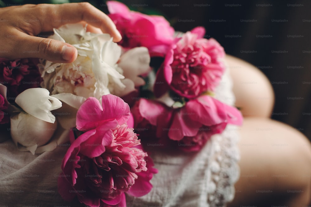 boho girl in white bohemia dress holding beautiful pink peonies on legs, top view. space for text. stylish hipster woman with flower in hand. moody morning. atmospheric sensual moment
