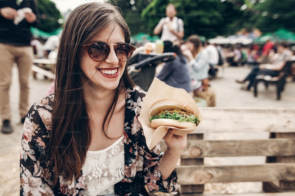 stylish hipster woman in sunglasses with red lips holding juicy burger. boho girl holding  hamburger and smiling at street food festival. summertime. summer vacation travel