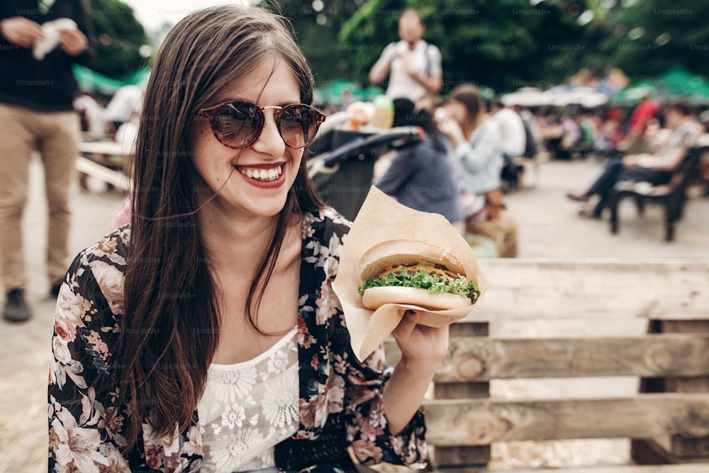 빨간 입술을 가진 선글라스를 쓴 세련된 힙스터 여자는 육즙이 많은 버거를 들고 있다. 햄버거를 들고 길거리 음식 축제에서 웃고 있는 보호 소녀. 여름철. 여름 방학 여행