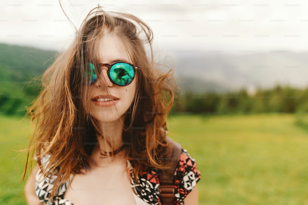 stylish hipster woman traveler with fashionable sunglasses and windy hair with confident look posing on top of mountains in summer, travel concept, space for text