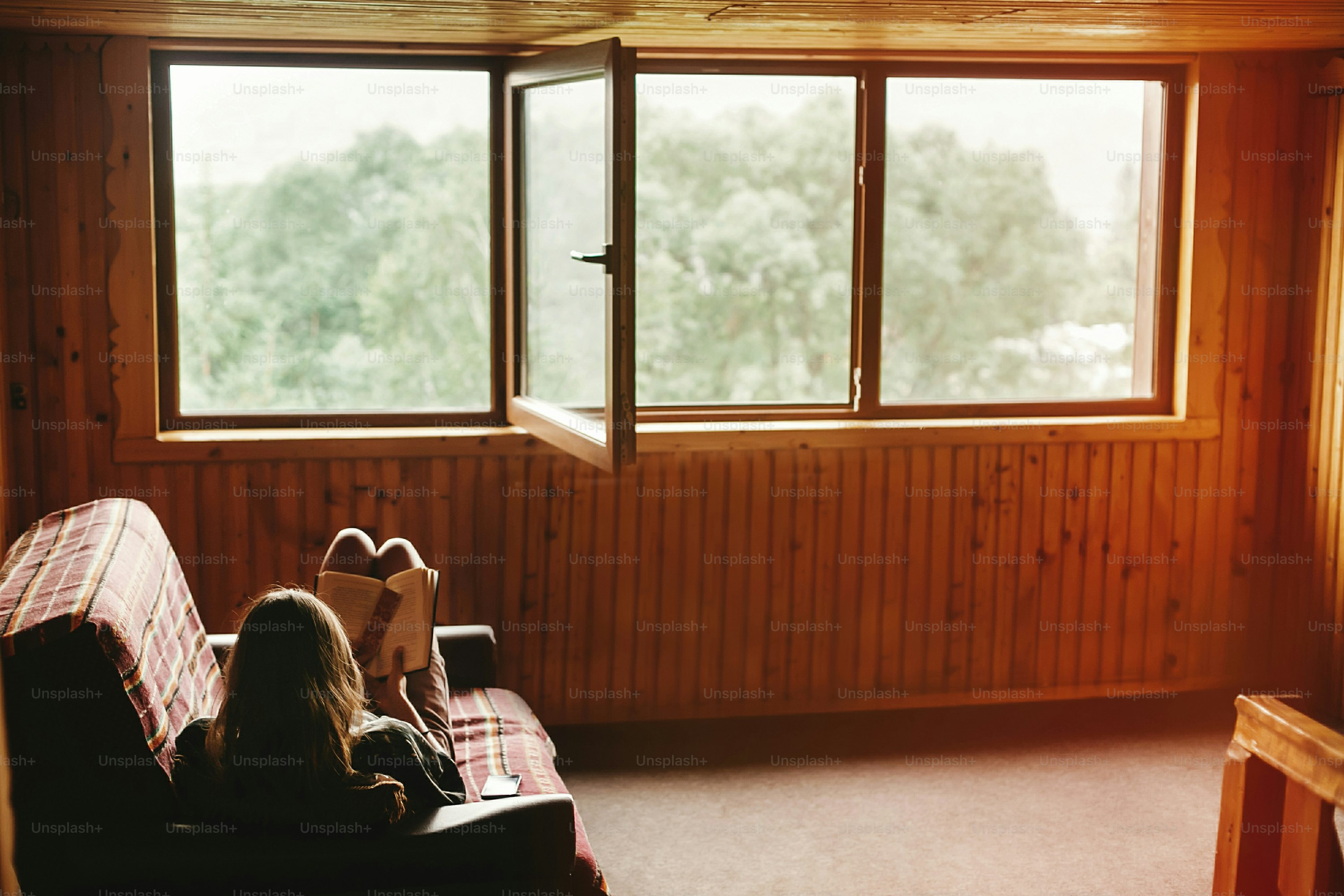 hipster woman sitting on coach and reading book in wooden cottage with view on woods, summer vacation concept, space for text