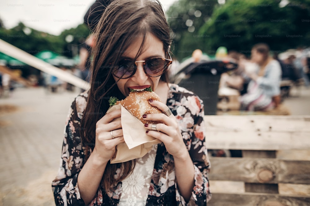 Mujer hipster con estilo sosteniendo una jugosa hamburguesa y comiendo. Chica boho mordiendo hamburguesa sonriendo en el festival de comida callejera. verano. Viajes de vacaciones de verano. Espacio para el texto