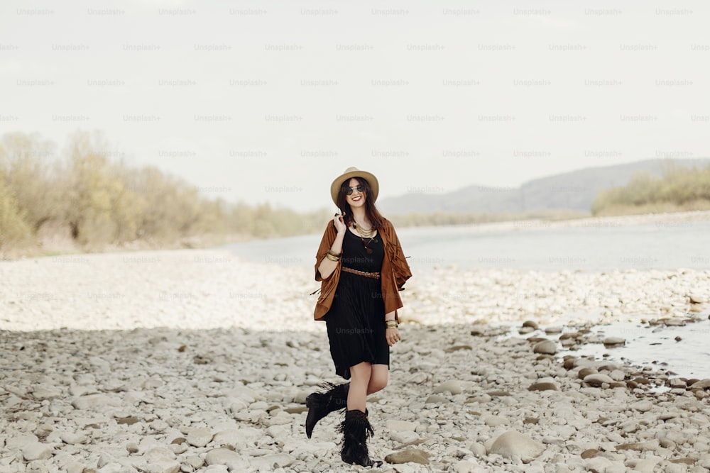 stylish boho traveler woman in hat, fringe poncho walking near water river beach in mountains, gypsy hipster girl. wanderlust summer travel. atmospheric moment. space for text.