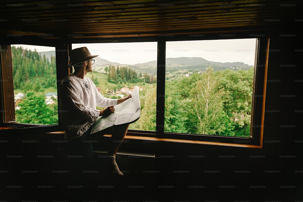 stylish hipster man sitting at window with view on mountains and holding map and exploring in wooden house, summer travel concept, space for text