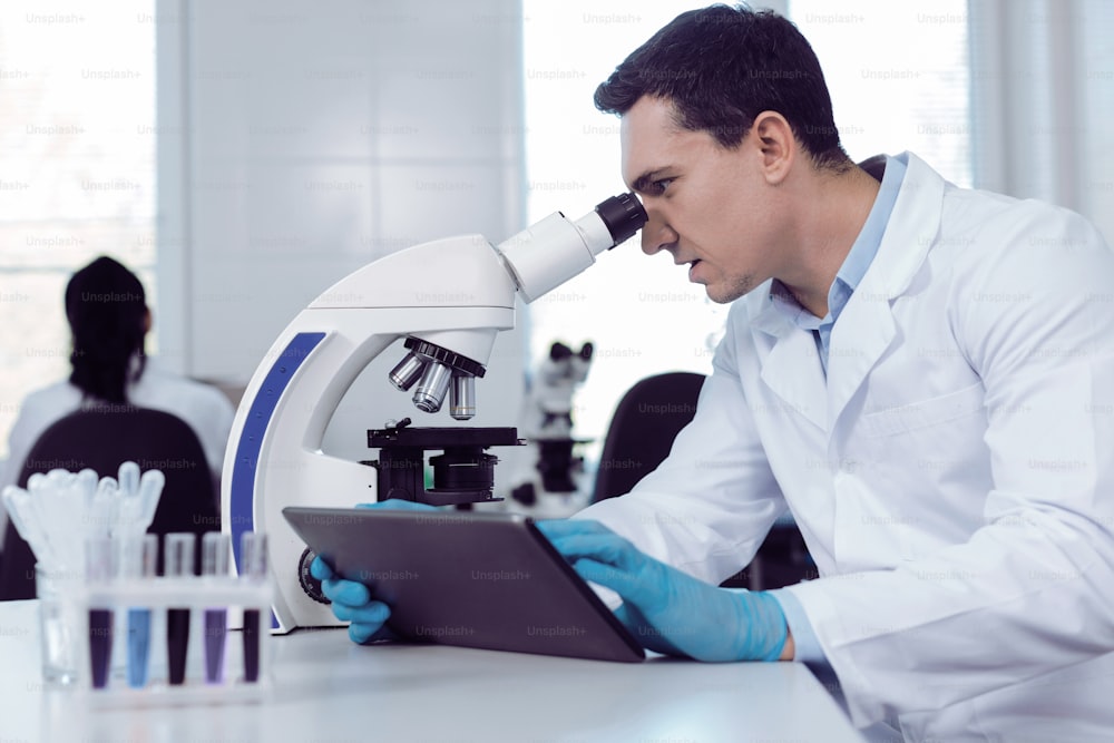En el laboratorio. Científico masculino inteligente positivo sosteniendo una tableta y mirando al microscopio mientras trabaja en el laboratorio biológico