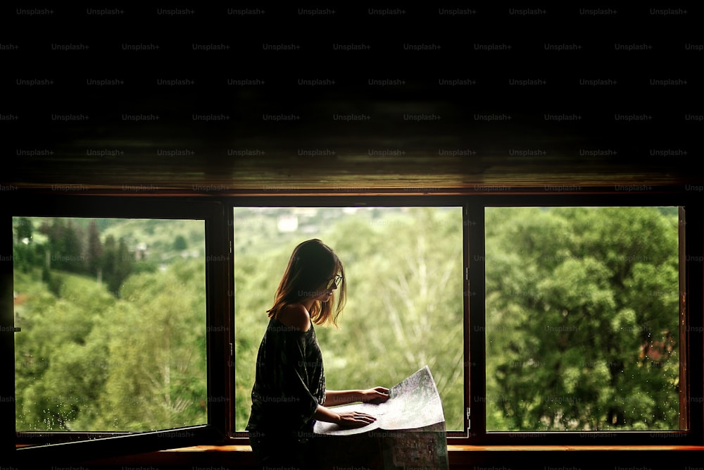 Mujer sosteniendo mapa sentada en la ventana con vista a las montañas asombrosas en casa de madera, planificación de viajes de verano o concepto de arquitecto, espacio para texto
