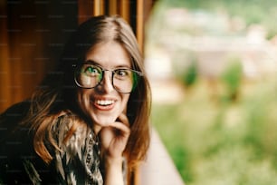 stylish hipster woman sitting at window with amused smile  in wooden house, funny moment, summer travel concept, space for text