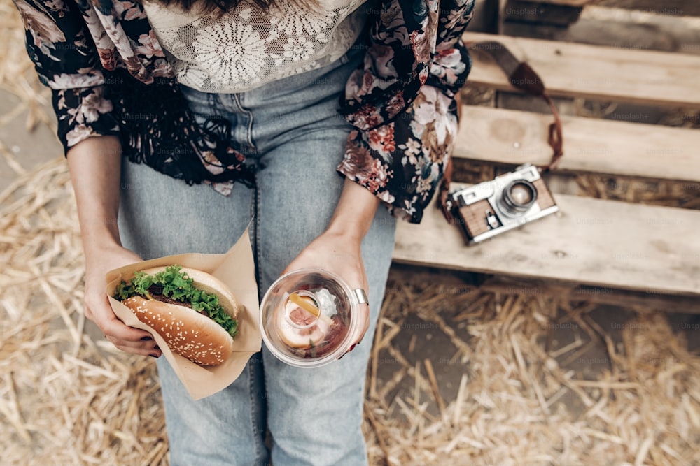 juicy burger and lemonade in hands, top view. stylish hipster woman holding  cheeseburger and refreshing drink. boho girl at street food festival. summer vacation picnic. space for text