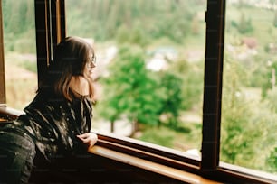 stylish hipster woman  sitting at window with view at amazing mountains in wooden house, summer travel concept, space for text
