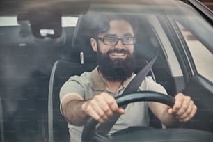 Um jovem atraente dirigindo um veículo, olhando para a paisagem, visto através do vidro do para-brisa. Motorista feliz, de mãos dadas no volante com um largo sorriso bonito.