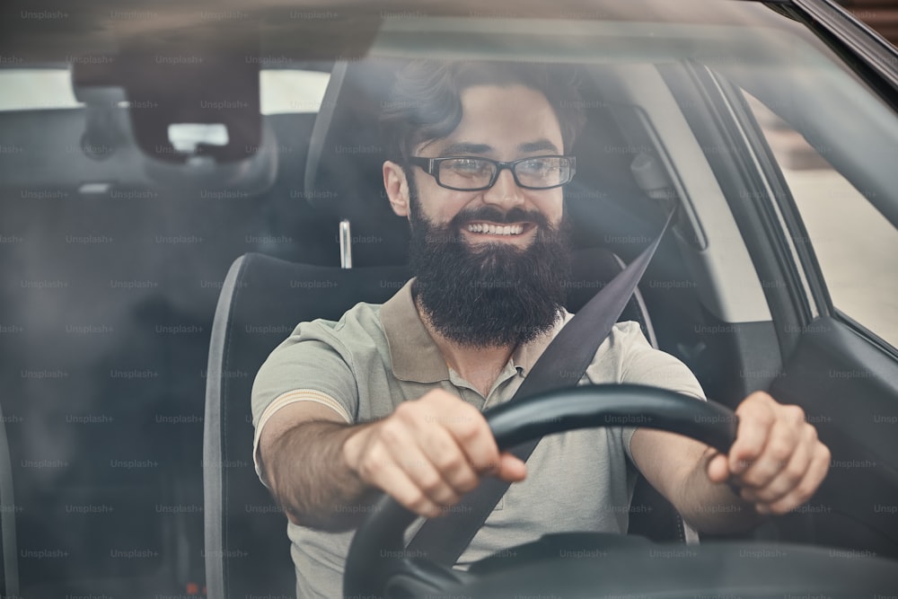 Un joven atractivo que conduce un vehículo, mirando el paisaje, visto a través del vidrio del parabrisas. Conductor feliz, tomado de la mano en el volante con una amplia y hermosa sonrisa.