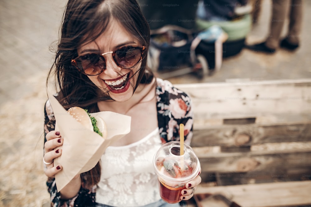 Stilvolle Hipster-Frau mit saftigem Burger und Limonade beim Essen. Boho-Mädchen beißt Hamburger, hat Spaß, lächelt beim Street Food Festival. Sommerzeit. Sommerferien Picknick. Platz für Text