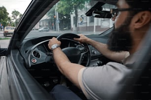 Angled side view modern casual bearded man, wearing glasses, watch and T-shirt, driving the car. Close up drivers' hands.