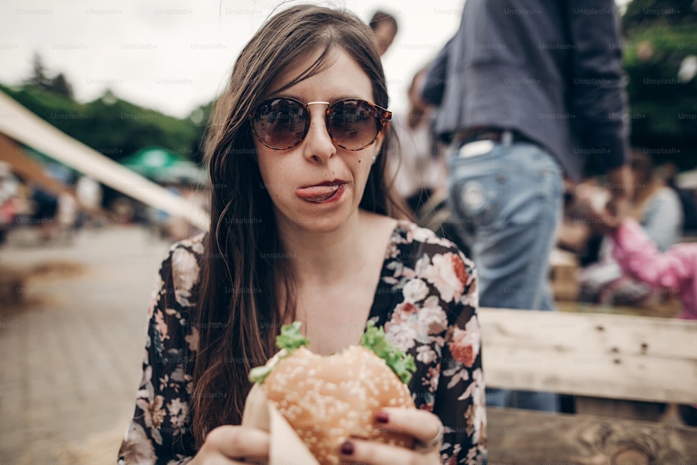 ジューシーなハンバーガーを食べるスタイリッシュなヒップスターの女性。おいしいチーズバーガーを噛む自由奔放に生きる女の子、屋台の食べ物の祭りで笑顔。夏時。夏休みの旅行ピクニック。テキスト用のスペース