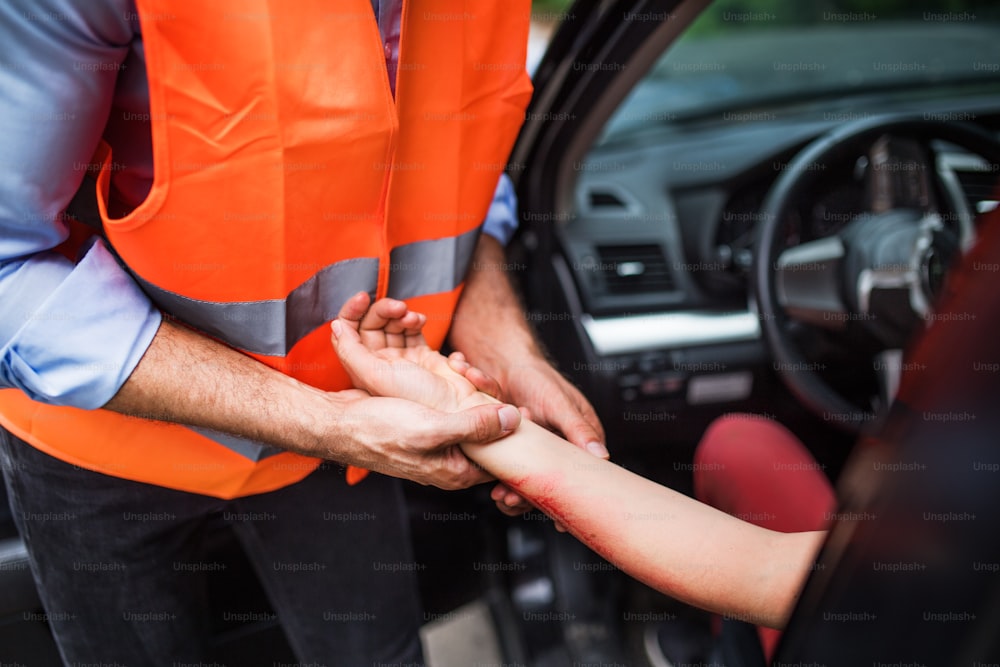 주황색 반사 조끼를 입은 남자가 여성 운전자의 맥박을 확인하고 있다. 교통사고 후 차에 앉아 있는 젊은 여성의 팔.