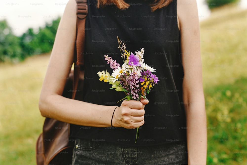 Mujer hipster con estilo con mochila que recoge y sostiene flores silvestres en la mano en las montañas de verano, concepto de viaje