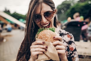 ジューシーなハンバーガーを食べるスタイリッシュなヒップスターの女性。チーズバーガーを噛む自由奔放に生きる女の子、屋台の食べ物の祭典で笑顔。夏時。夏休みの旅行ピクニック。テキスト用のスペース