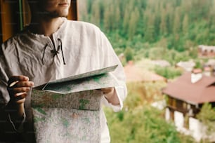 stylish hipster man sitting at window with view on mountains and holding map and making route for hiking in wooden house, summer travel concept, space for text