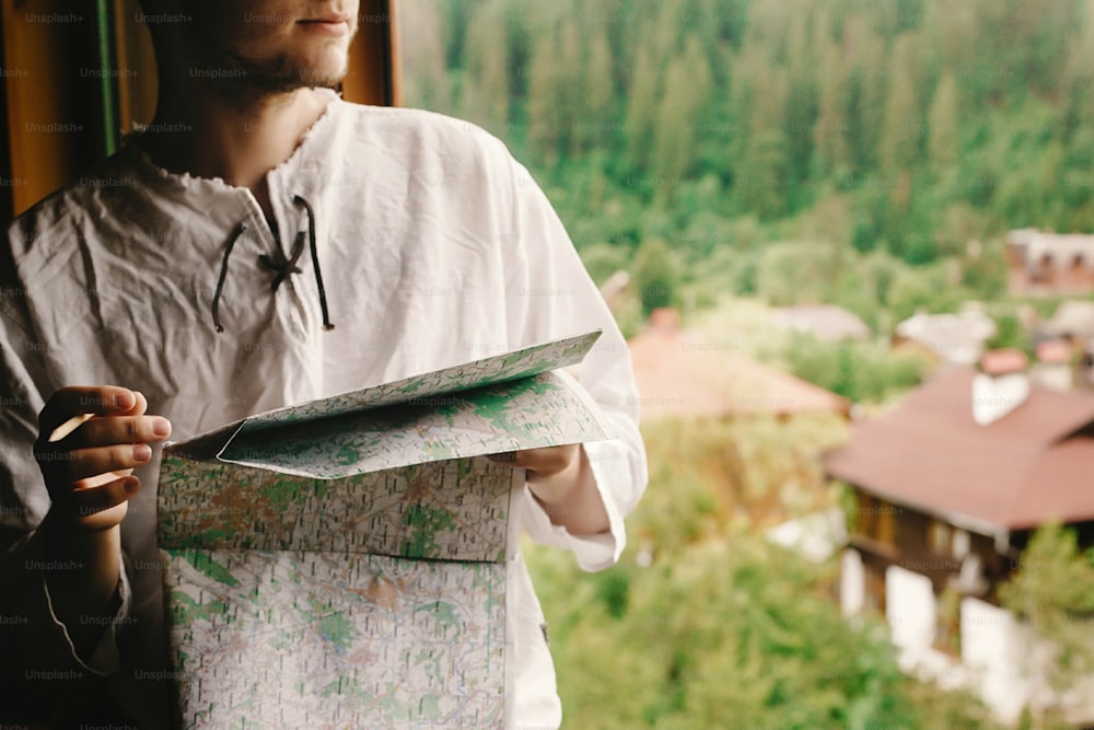 Stylischer Hipster-Mann sitzt am Fenster mit Blick auf die Berge und hält Karte und macht Route zum Wandern im Holzhaus, Sommerreisekonzept, Platz für Text