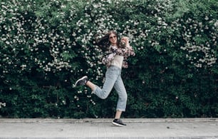 happy hipster woman in sunglasses having fun on background of blooming bush with white flowers of spirea. boho girl jumping and smiling in modern clothes in summer street. space for text