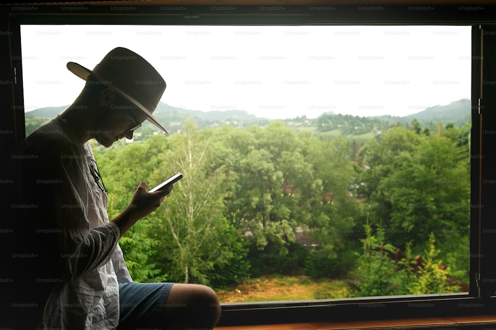 힙스터 남자는 전화를 들고 그것을 보고, 산, 여행 또는 통신 탐색 개념, 텍스트 공간에 대한 전망이 있는 목조 주택의 창문에 앉아