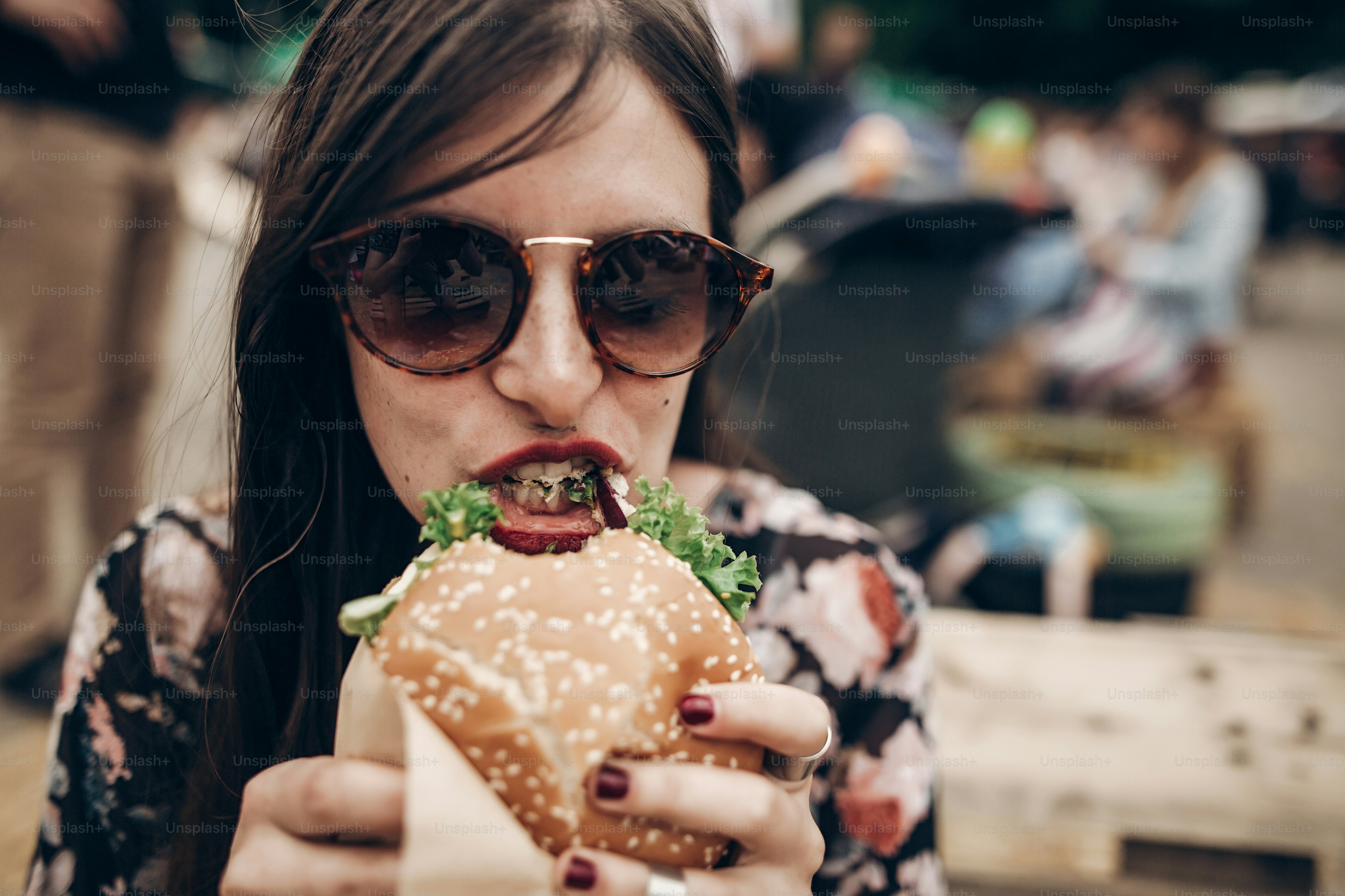 A woman wearing sunglasses eating a large burger with lettuce at an outdoor event