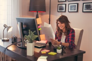 Young woman sitting at a desk at her home office, holding documents joyful after a business success