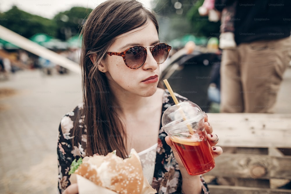 Mujer hipster con estilo bebiendo limonada. Chica boho genial en denim y ropa bohemia, con cóctel y hamburguesa en el festival de comida callejera. verano. Picnic de vacaciones de verano
