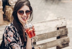 Stylische Hipster-Frau mit Sonnenbrille und roten Lippen trinkt Limonade. Boho-Mädchen hält Cocktail und lächelt beim Street Food Festival. Sommerzeit. Sommerferien reisen. Platz für Text