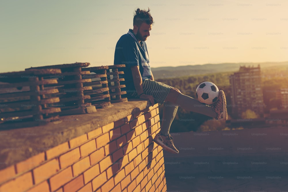 Jogador de futebol em um telhado de edifício, sentado e relaxando após a partida, segurando uma bola