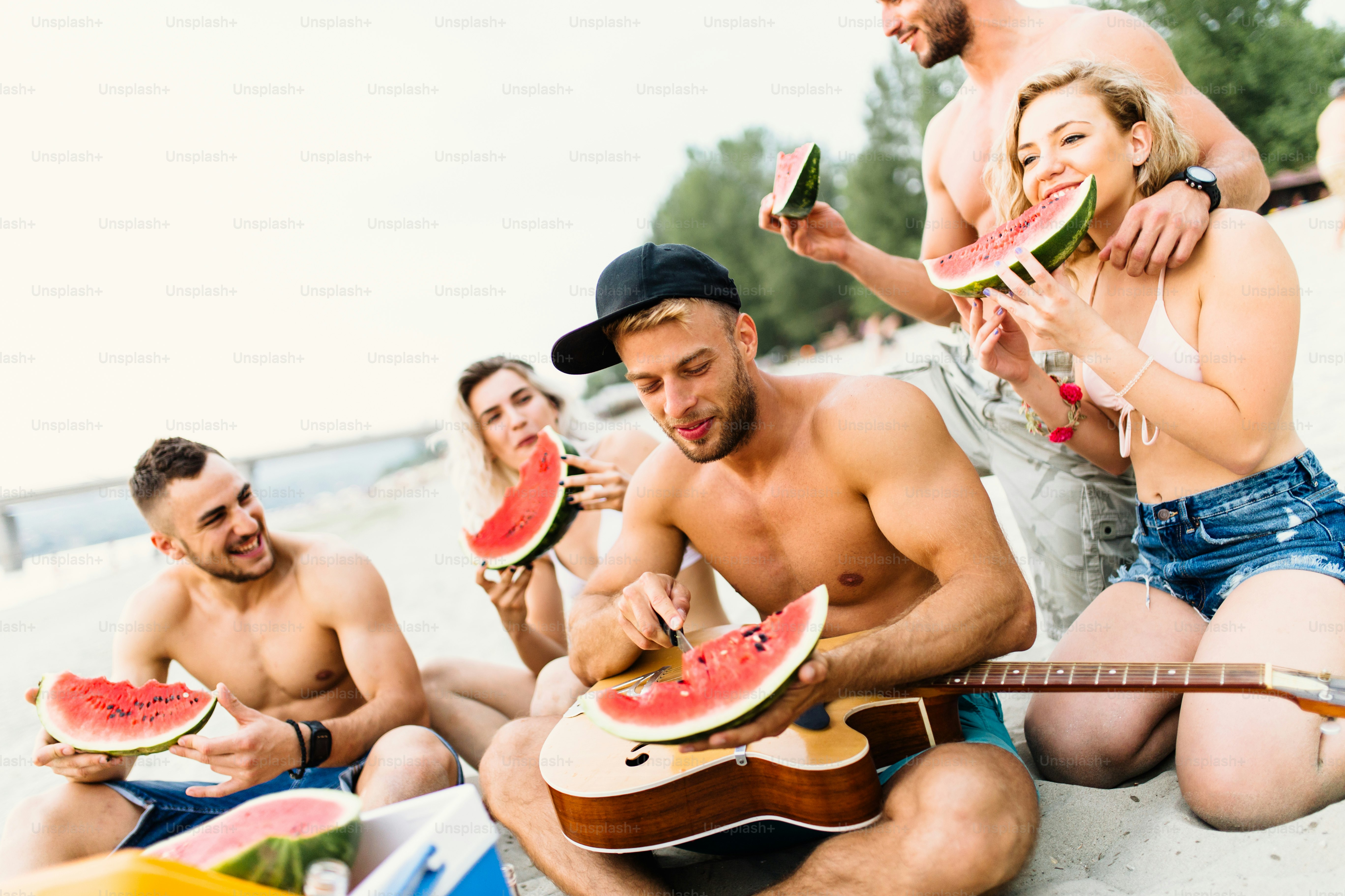 Gruppe attraktiver junger Leute, die am Strand Wassermelone trinken und essen.