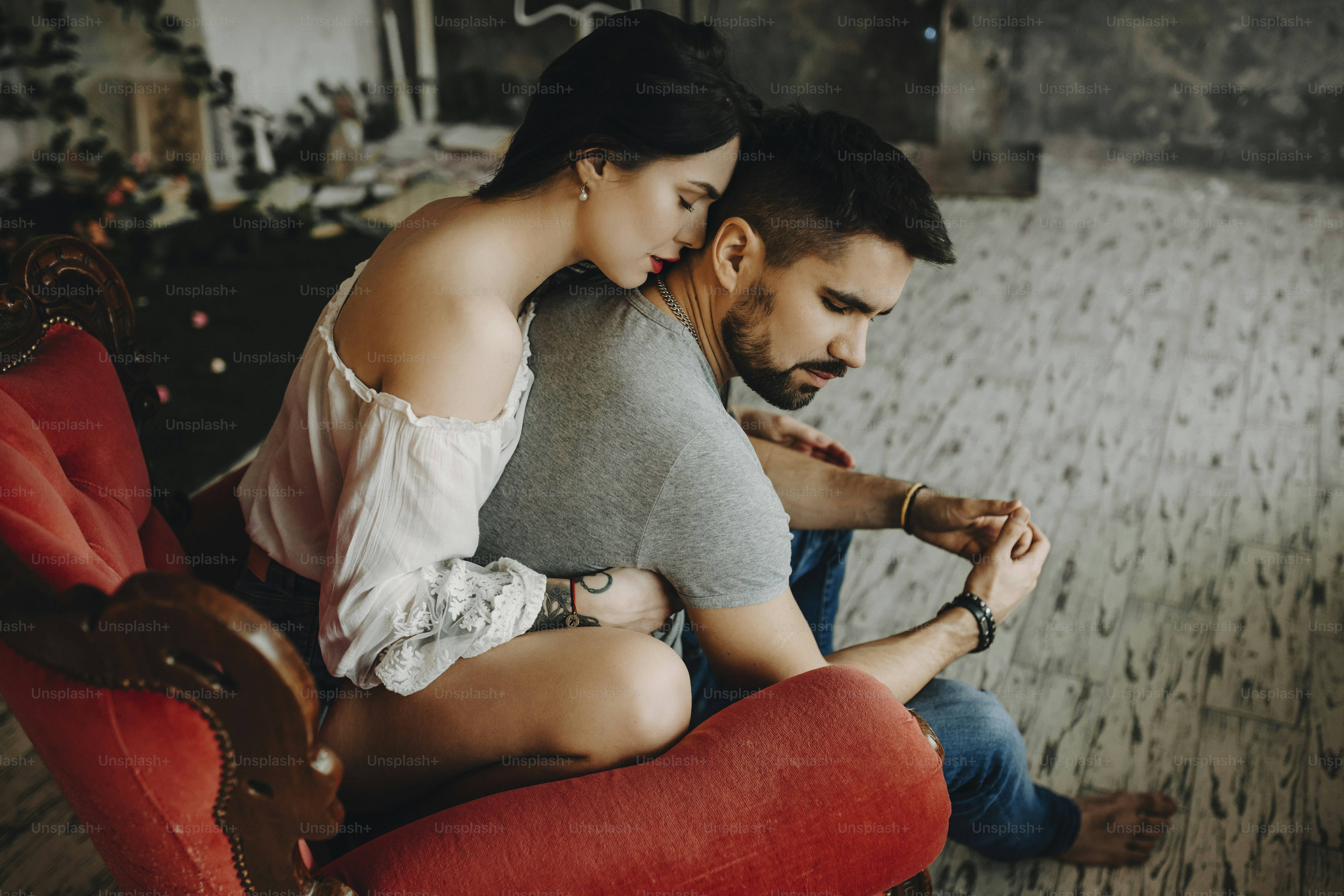 Very beautiful couple sitting on vintage red chair, where tattooed with red lips girl is embracing from back his lover waiting to kiss his neck while he is turning his head to her gently.