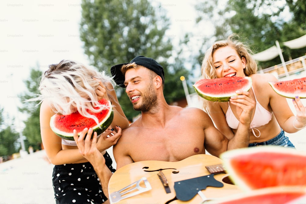 Gruppo di giovani attraenti che si divertono in spiaggia, bevendo e mangiando anguria.