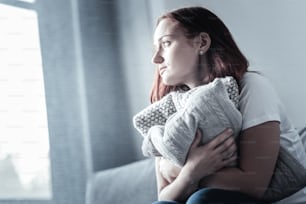 His presence. Depressed pleasant lamentable woman posing with pillow while turning face in profile and sitting on blurred background